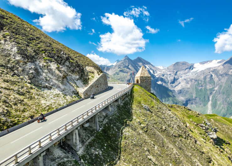 Motorrijders rijden over de Grossglockner Hochalpenstrasse, slingerend langs stenen muren en besneeuwde bergtoppen onder een blauwe hemel.