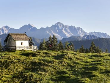 Twee fietsers rijden langs een kleine kapel in Saalfelden-Leogang, met bergen onder een heldere hemel.