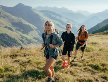 Drie vrouwen wandelen op een zonnige dag over een grassig bergpad in Saalbach-Hinterglemm met schilderachtige bergtoppen.