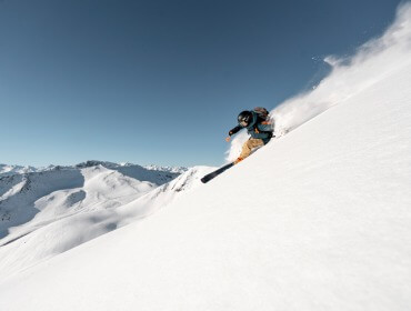 Een skiër in zijn versnelling scheurt over een steile helling van Saalbach-Hinterglemm, terwijl hij poeder opspuit onder een strakblauwe hemel.