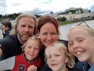 Family in Salzburg Een lachend gezin van vijf poseert voor een selfie bij een rivier met een schilderachtig Europees stadje op de achtergrond.