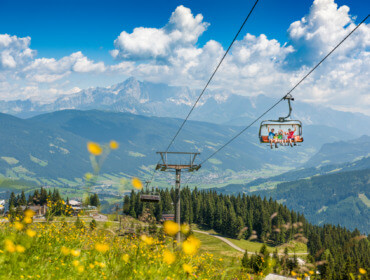 160720-flachau-dsc_1983 Mensen rijden met een skilift over de groene bergen van Flachau, wilde bloemen bloeien onder een helderblauwe hemel.