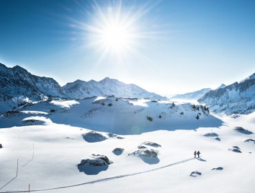 Twee mensen wandelen op een besneeuwd bergpad in Obertauern, de zon schijnt boven de besneeuwde toppen.