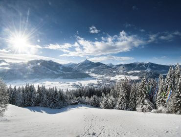 2016_Altenmarkt_Ort_©Huber- Besneeuwd landschap in Altenmarkt-Zauchensee met dennenbomen, bergen in de verte en een felle zon aan een helderblauwe hemel.