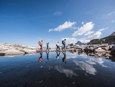 Vier mensen wandelen over rotsachtig terrein naast Uttendorf Weißsee, een helder meer dat lucht en wolken weerspiegelt onder een blauwe hemel.