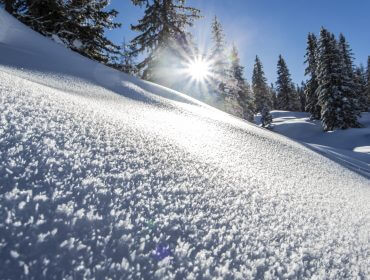 Zon schijnt over besneeuwde heuvel met ijzige bomen op de achtergrond onder een strakblauwe lucht.