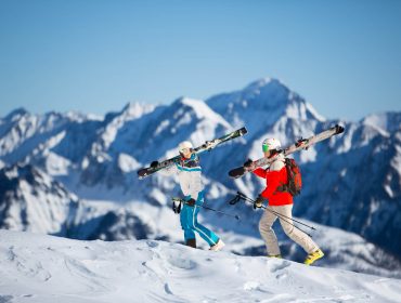 Twee skiërs met ski's lopen op de besneeuwde berg Salzburger Lungau met bergtoppen op de achtergrond onder een heldere hemel.