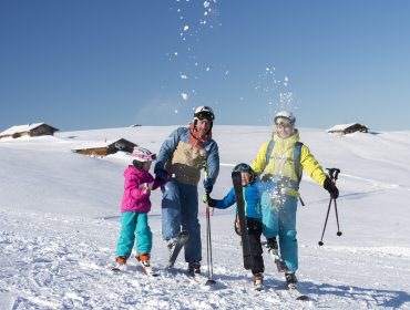 Een gezin van vier in kleurrijke skikleding spelend in het Salzburger Saalachtal met besneeuwde heuvels en hutten achter hen.