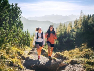 Twee vrouwen wandelen op een zonnig bergpad, omringd door bomen en verre bergtoppen.