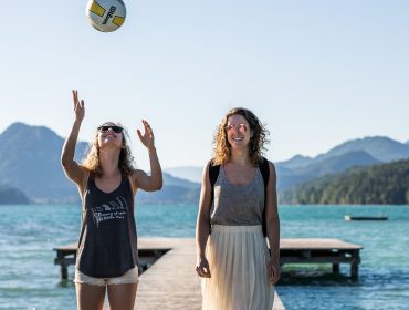 2017-06-19-slt-sommershooting-by-michael-groessinger-img_0304-e1500535974669 Twee vrouwen op een steiger bij een meer, één gooit een volleybal, bergen en blauwe lucht op de achtergrond.