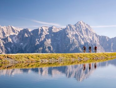 asitz-bike-by-klemens-koenig_highres-38-e1649930243402 Twee fietsers rijden over een graspad langs een meer met hoge bergen op de achtergrond.