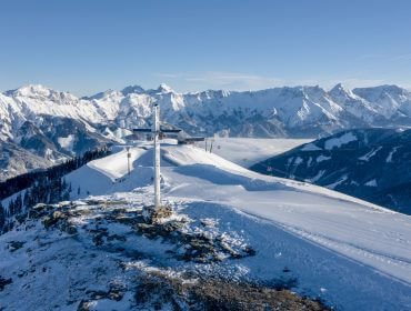 asitz-by-lemonmedia-2 Besneeuwde bergtop met een kruis, uitkijkend over een reeks besneeuwde toppen onder een strakblauwe hemel.