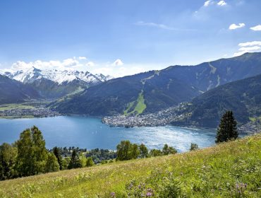 Een schilderachtig meer omringd door bergen, een klein stadje en met gras begroeide heuvels onder een strakblauwe hemel.