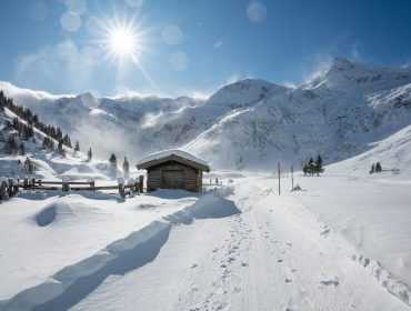 Een kleine houten hut in een besneeuwd berglandschap onder een stralende zon en een helderblauwe hemel.