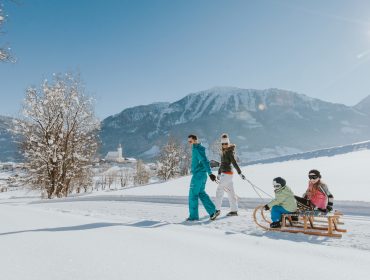 Twee volwassenen trekken twee kinderen op een slee door een besneeuwd landschap met bergen op de achtergrond.