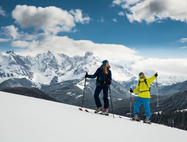 Twee mensen skiën bergop op een besneeuwde helling met besneeuwde bergen en wolken op de achtergrond.
