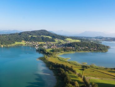 Luchtfoto van een dorp aan een meer met groene heuvels en bergen in de verte onder een strakblauwe lucht.