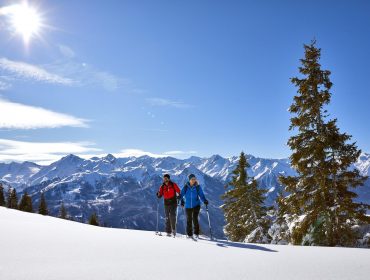Twee mensen langlaufen op een besneeuwde berg met dennenbomen en een felle zon erboven.