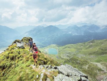 Drie wandelaars lopen over een met gras begroeide bergrug met een schilderachtig uitzicht op valleien, een meer en bergen in de verte.