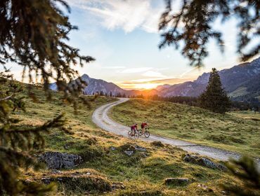 Twee fietsers rijden over een kronkelend bergpad bij zonsondergang, omlijst door bomen en bergtoppen in de verte.