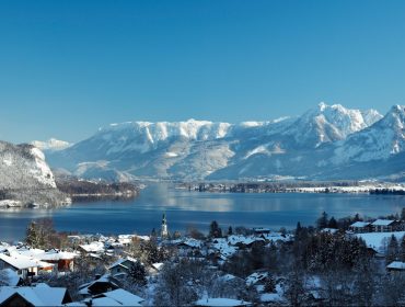 Besneeuwd österreichisch dorp aan een meer, omringd door bergen onder een strakblauwe hemel.