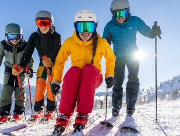 Vier skiërs in kleurrijke kleding glimlachen en poseren samen op een besneeuwde helling onder een heldere, zonnige hemel.