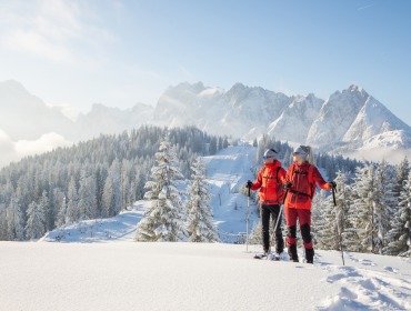 Dachstein West Skitour-237 Twee skiërs in rode outfits staan op een besneeuwde heuvel, met uitzicht op pijnbomen en besneeuwde bergen.