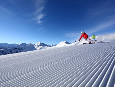 Twee skiërs glijden over een vers geprepareerde helling met besneeuwde bergen en een helderblauwe lucht op de achtergrond.