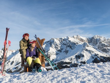 Twee skiërs zitten op een bankje in besneeuwde bergen, met skispullen en bergtoppen op de achtergrond onder een heldere hemel.