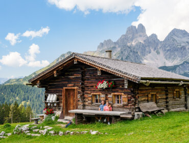 Een rustieke houten hut in de bergen met twee mensen die buiten op een bankje zitten, omgeven door groen.