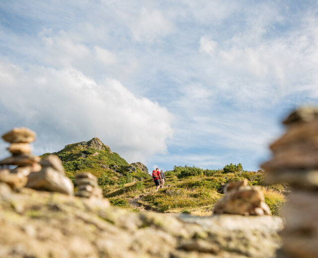 Twee wandelaars lopen over een bergpad met stenen stèles op de voorgrond en een bergtop op de achtergrond.