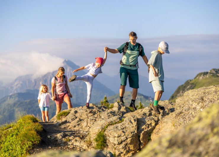 Vier kinderen en een volwassene wandelen over rotsachtig terrein met bergen op de achtergrond onder een heldere hemel.
