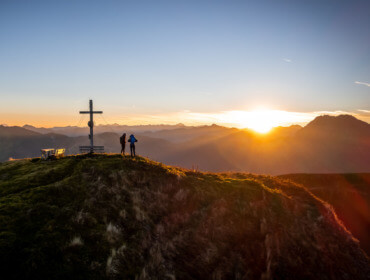 Twee mensen staan bij een kruis op een berg bij zonsopgang, met uitzicht op bergtoppen in de verte.