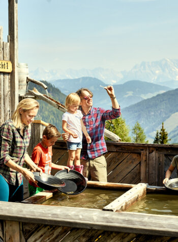 Een familie zoekt goud bij een houten trog met bergen en bossen op de achtergrond.