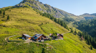 Houten hutten staan op een groene heuvel met een bergtop en bomen op de achtergrond onder een heldere hemel.