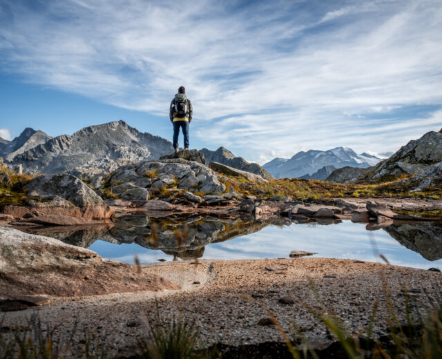 ©TVB Grossarltal; Weinschnabel-Albert-Biwak-Mureck-Arlscharte-Schoedersee Een wandelaar staat op een rotsachtige heuveltop met uitzicht op bergen, weerspiegeld in een heldere vijver onder een blauwe hemel.