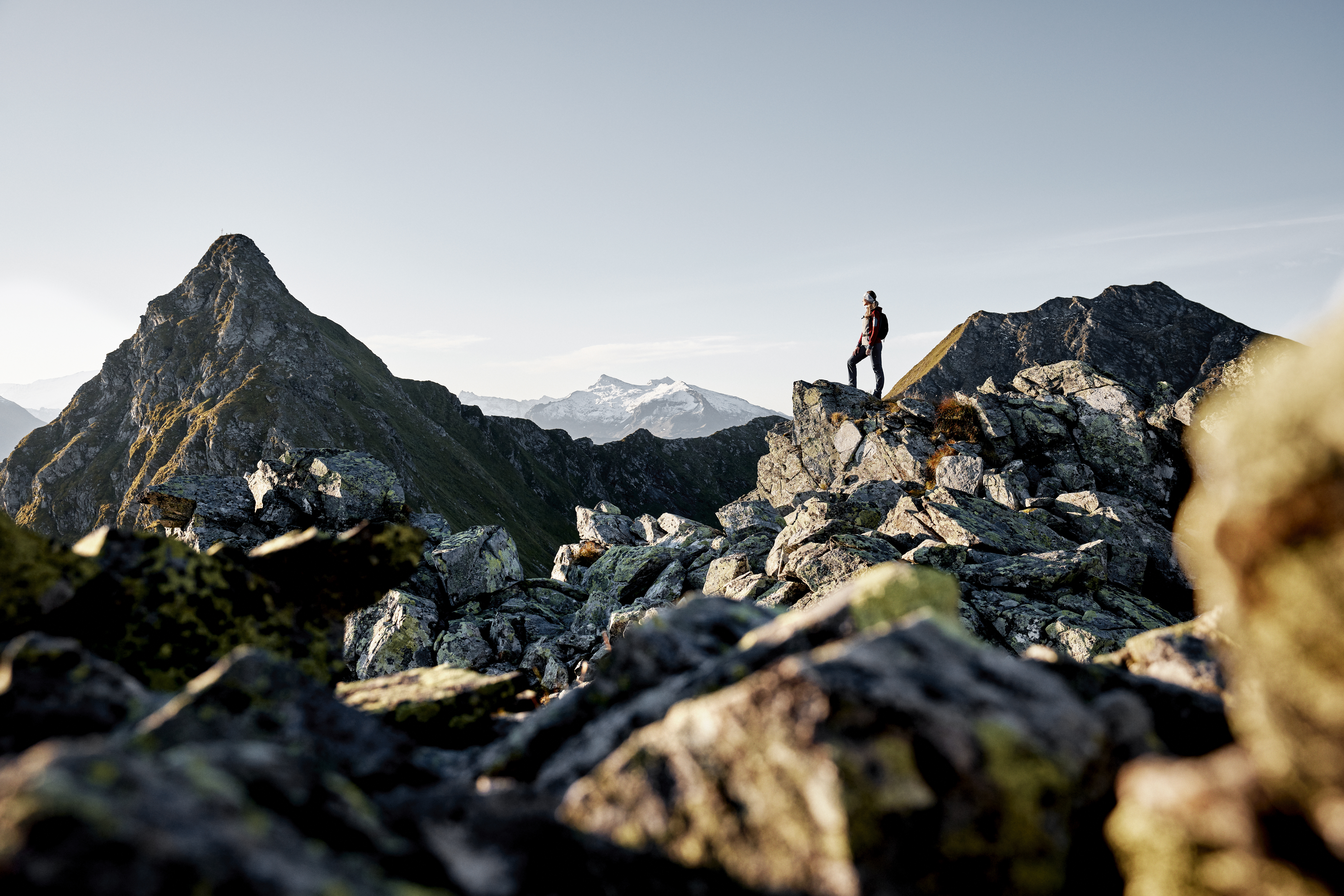 Een eenzame wandelaar staat op rotsachtig bergterrein, met bergtoppen en een heldere hemel op de achtergrond.