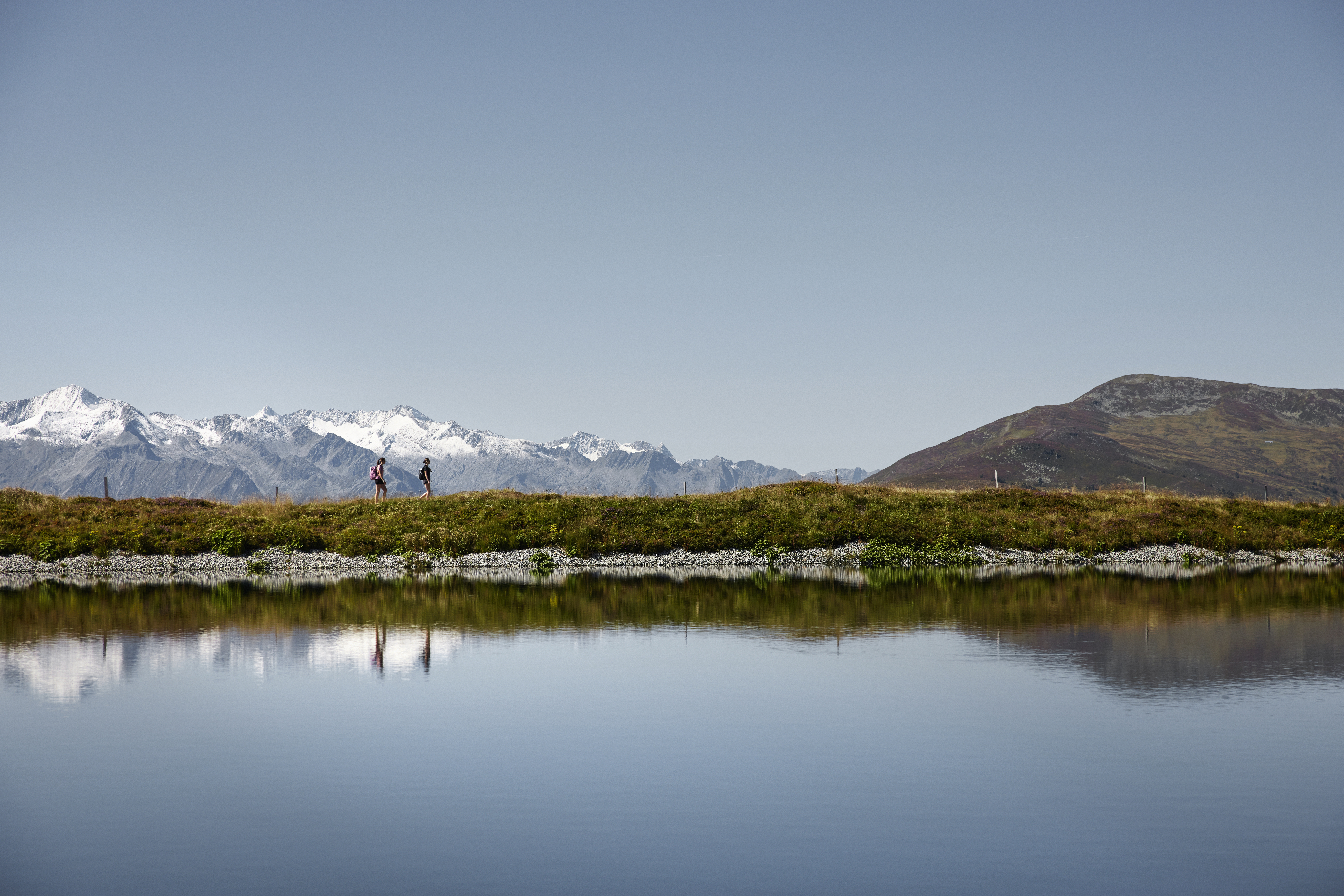 Twee mensen wandelen over een grasrand bij een rustig meer, met besneeuwde bergen op de achtergrond onder een heldere hemel.