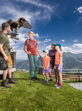 Een man toont een grote roofvogel aan een familie die buiten een rustiek houten gebouw op een berghelling staat.