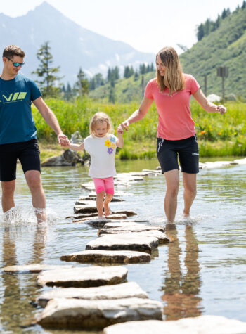 Een gezin van drie wandelt over stenen trappen over een ondiep beekje in een schilderachtig berglandschap.