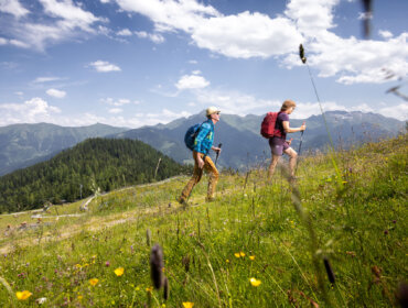 Sommershooting Rauriser Hochalmbahnen Twee mensen wandelen bergopwaarts door een grasland met bergen en blauwe lucht op de achtergrond.