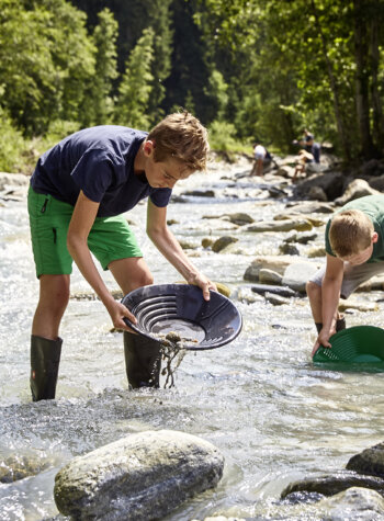 Twee jongens staan in een ondiepe rivier goud te zoeken met zwarte en groene pannen omringd door rotsen en bomen.