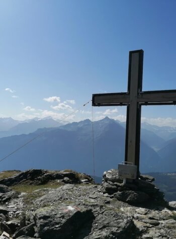 Een groot kruis staat bovenop een rotsachtige bergtop met bergketens in de verte onder een helderblauwe hemel.