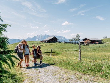 Een gezin wandelt op een zonnig pad in de buurt van houten hutten en bergen in een groen, grasachtig landschap.