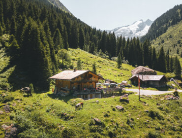 Een houten hut staat in een groene bergvallei met bomen en besneeuwde bergtoppen op de achtergrond.