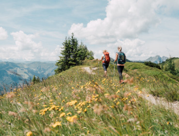Großarl Almenweg Twee mensen met rugzakken wandelen op een grassig bergpad omgeven door wilde bloemen onder een gedeeltelijk bewolkte hemel.