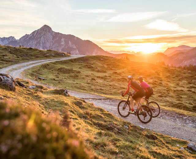 Gravelbike Twee fietsers rijden op een bergpad bij zonsondergang met schilderachtige bergtoppen op de achtergrond.