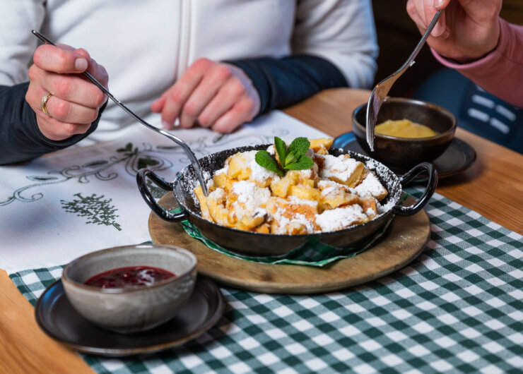 2023_Winter_Skifahren_Winter_Kulinarik_Kinder_Skifahren_Miriam_Lottes_(c)HochkoenigTourismusGmbH_web (54) Twee mensen delen een koekepan met een dessert met poedersuiker, jam en saus op een geruit tafelkleed.