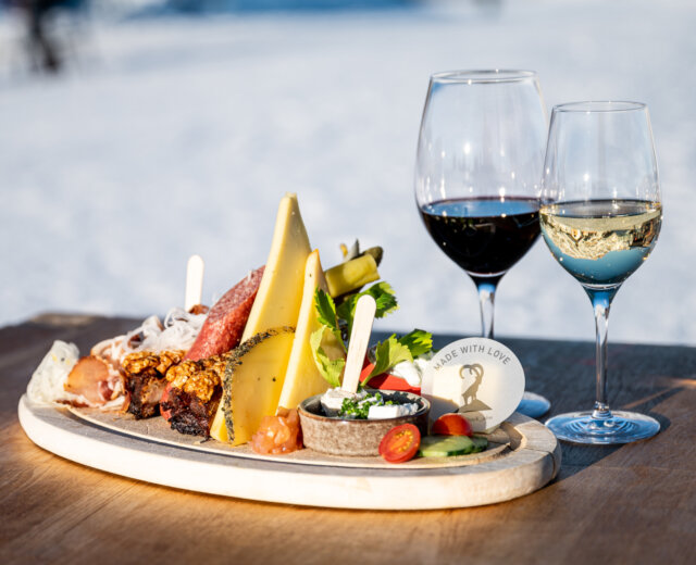 2023_Winter_Skifahren_Winter_Kulinarik_Kinder_Skifahren_Miriam_Lottes_(c)HochkoenigTourismusGmbH_web (67) Charcuterieplankje met kaas, vleeswaren en wijnglazen op een houten tafel buiten in het zonlicht.