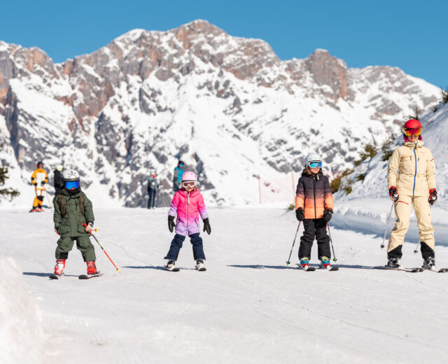 2023_Winter_Skifahren_Winter_Kulinarik_Kinder_Skifahren_Miriam_Lottes_(c)HochkoenigTourismusGmbH_web (93) Vier kinderen skiën op een besneeuwde helling met bergen op de achtergrond onder een strakblauwe hemel.